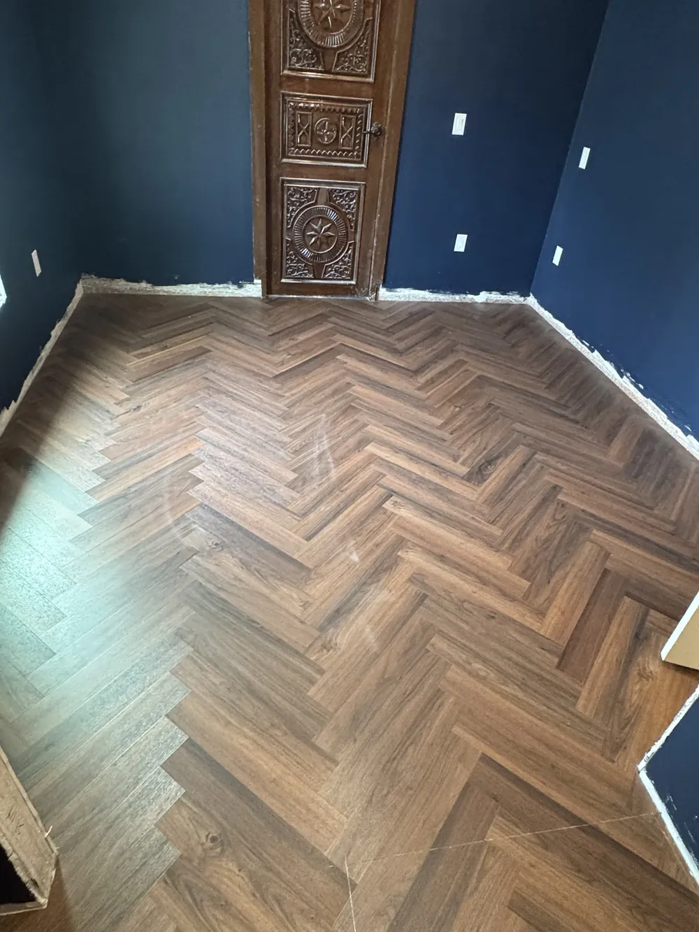Herringbone-pattern flooring installed up to a carved wood door in a navy-walled room